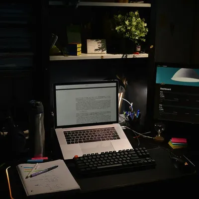 A laptop and notes on a desk in a dark room, a lamp on