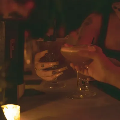 Two hands raising cocktails in a dim, candlelit bar